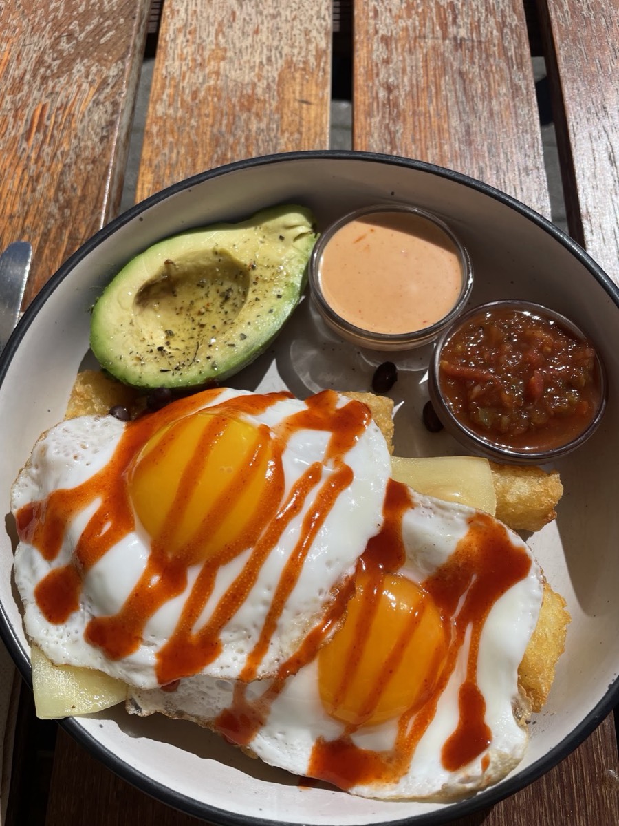 A plate with two fried eggs drizzled with orange sauce, half an avocado, cheese and two small pots of relish and sauce