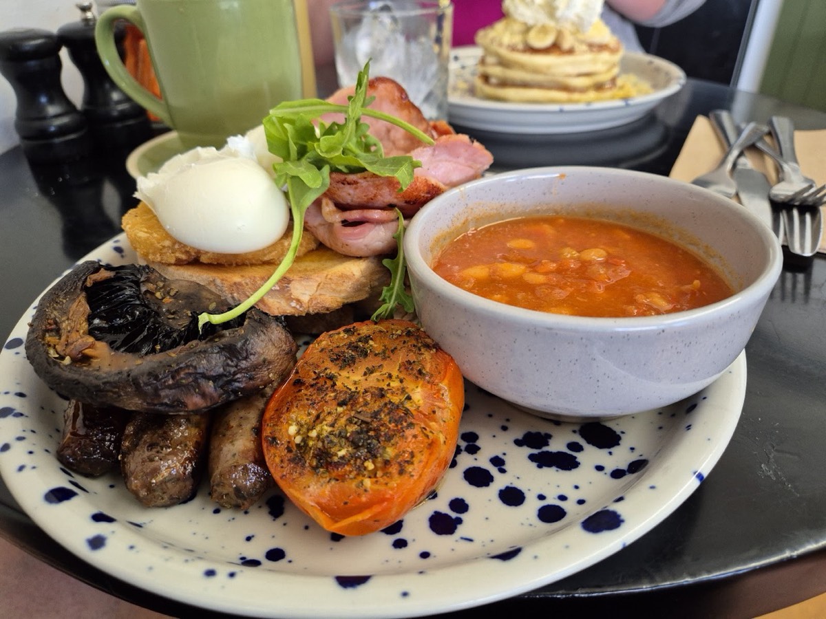 A big cooked breakfast on a speckled plate with a poached egg, bacon, sausages, mushroom and roast tomato, and a bowl of baked beans
