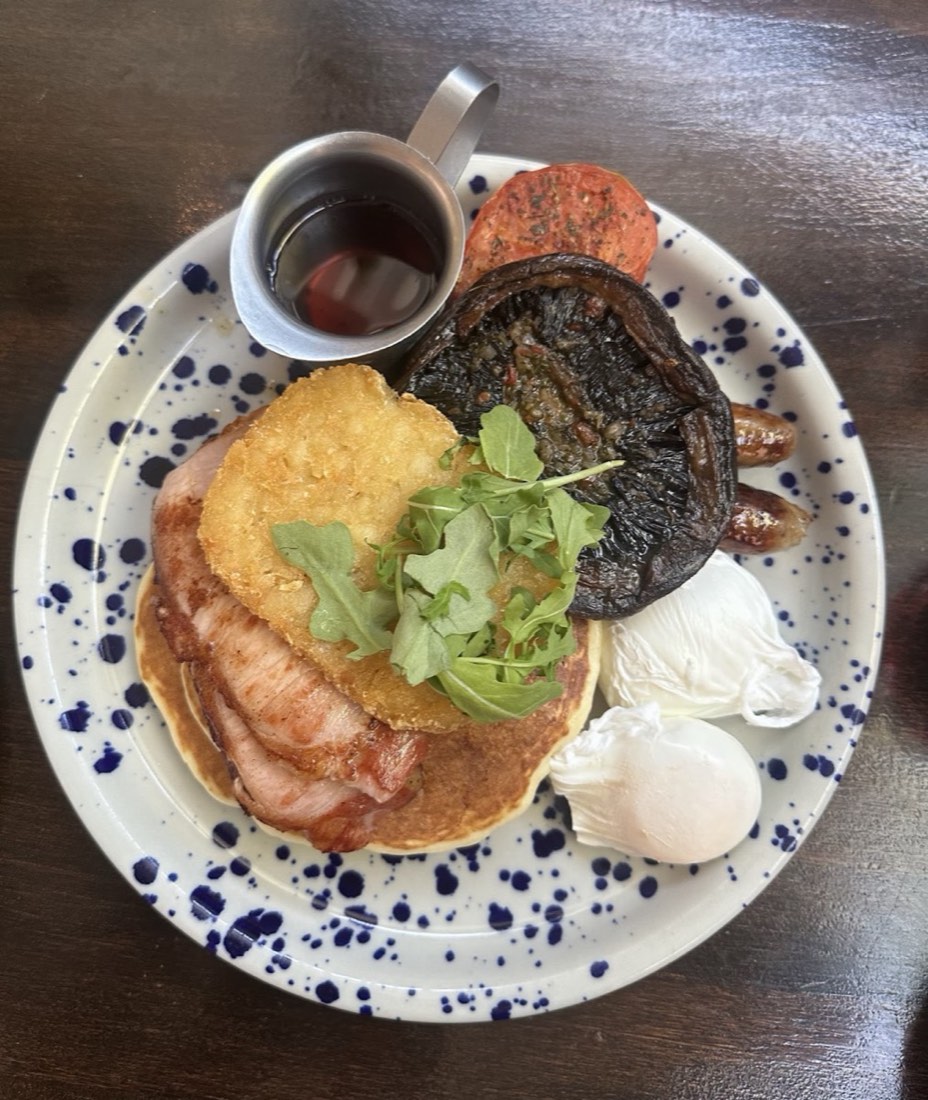 An overhead view of a speckled plate with a hash brown, bacon, rocket, two poached eggs, a mushroom and a small jug of sauce