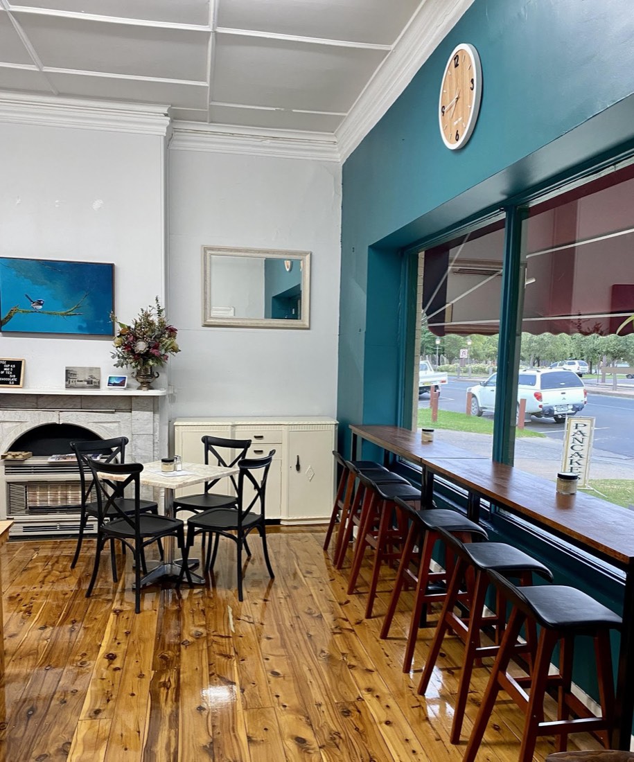 The dining room: a teal feature wall with a round clock and a big window, a long timber bench with bar stools, and a framed painting of a blue wren